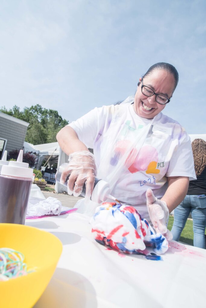 A woman wearing glasses and gloves smiles while squeezing dye onto a white T-shirt during an outdoor tie-dye activity. Bottles of dye and a yellow bowl with rubber bands are on the table.
