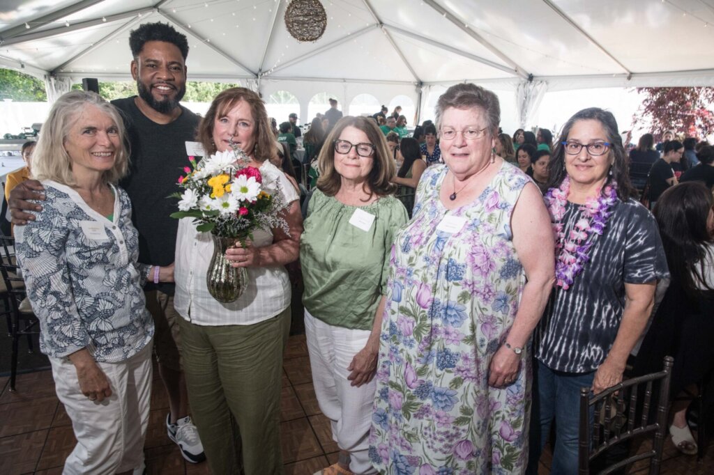 A group of six adults, five women and one man, stand together under a white event tent. One woman holds a bouquet of flowers. People are gathered at tables in the background. Everyone is smiling.