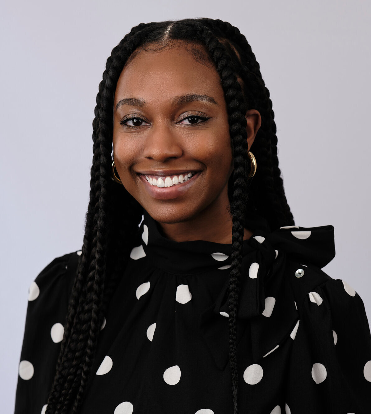 A woman with long, braided hair smiles at the camera. She wears gold hoop earrings and a black top with large white polka dots and a bow on the shoulder, set against a plain light gray background.
