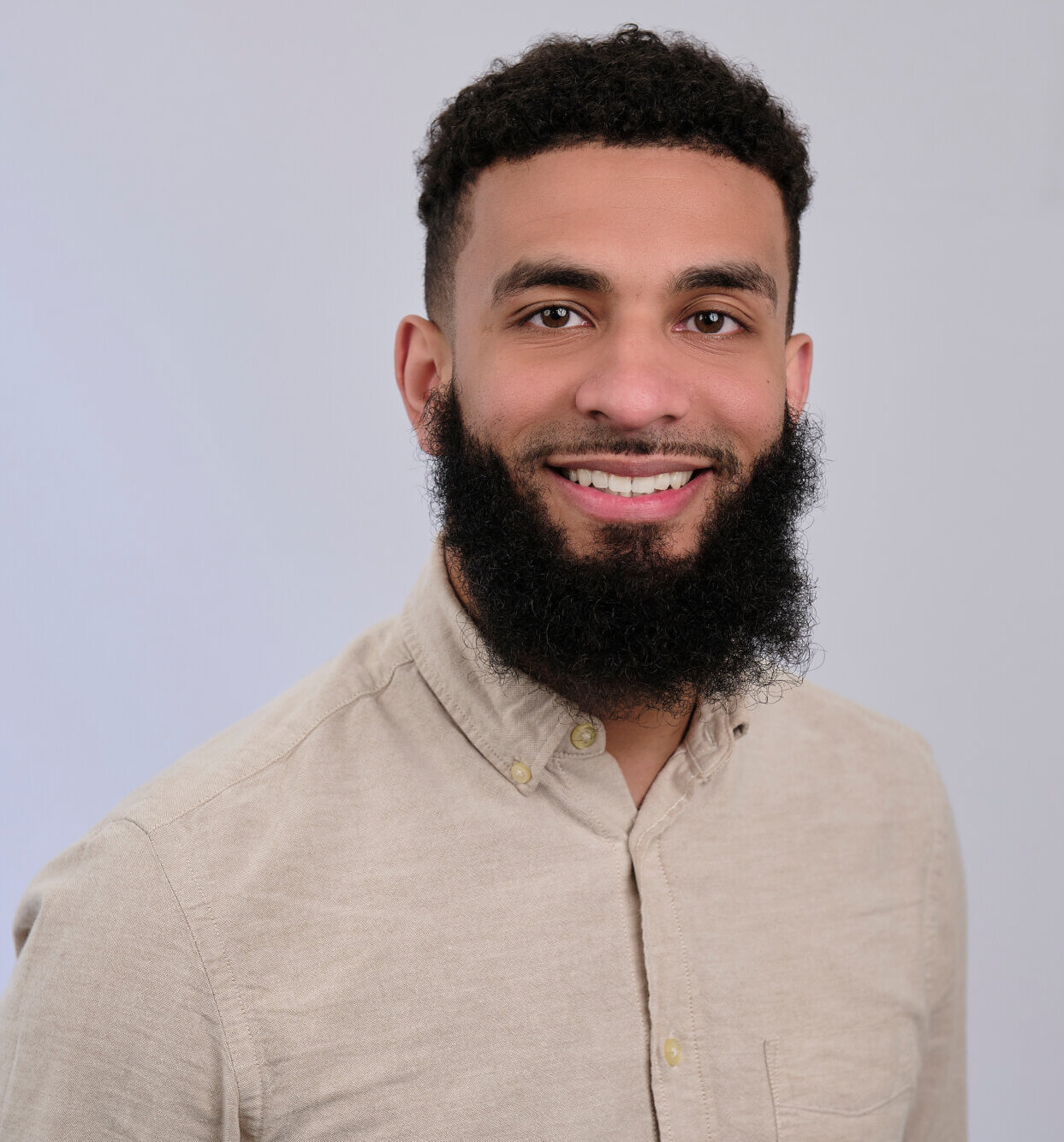 A man with curly hair and a full beard smiles at the camera. He is wearing a beige button-up shirt and is posed against a plain light gray background.