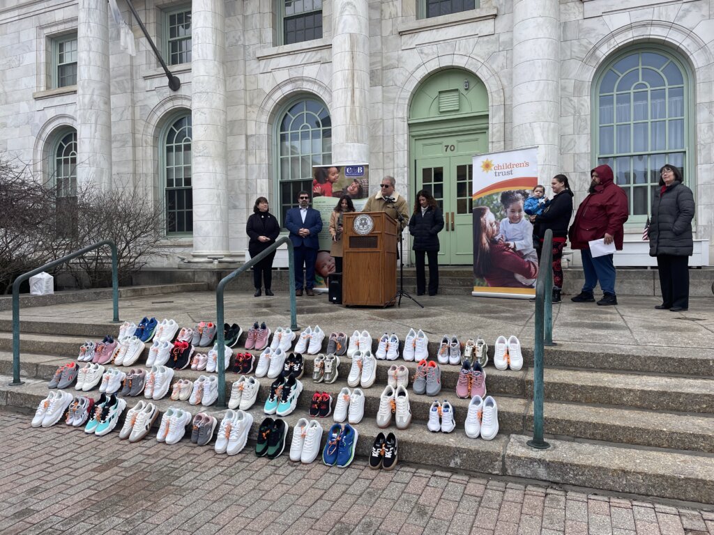 man speaking at podium with display of children's shoes