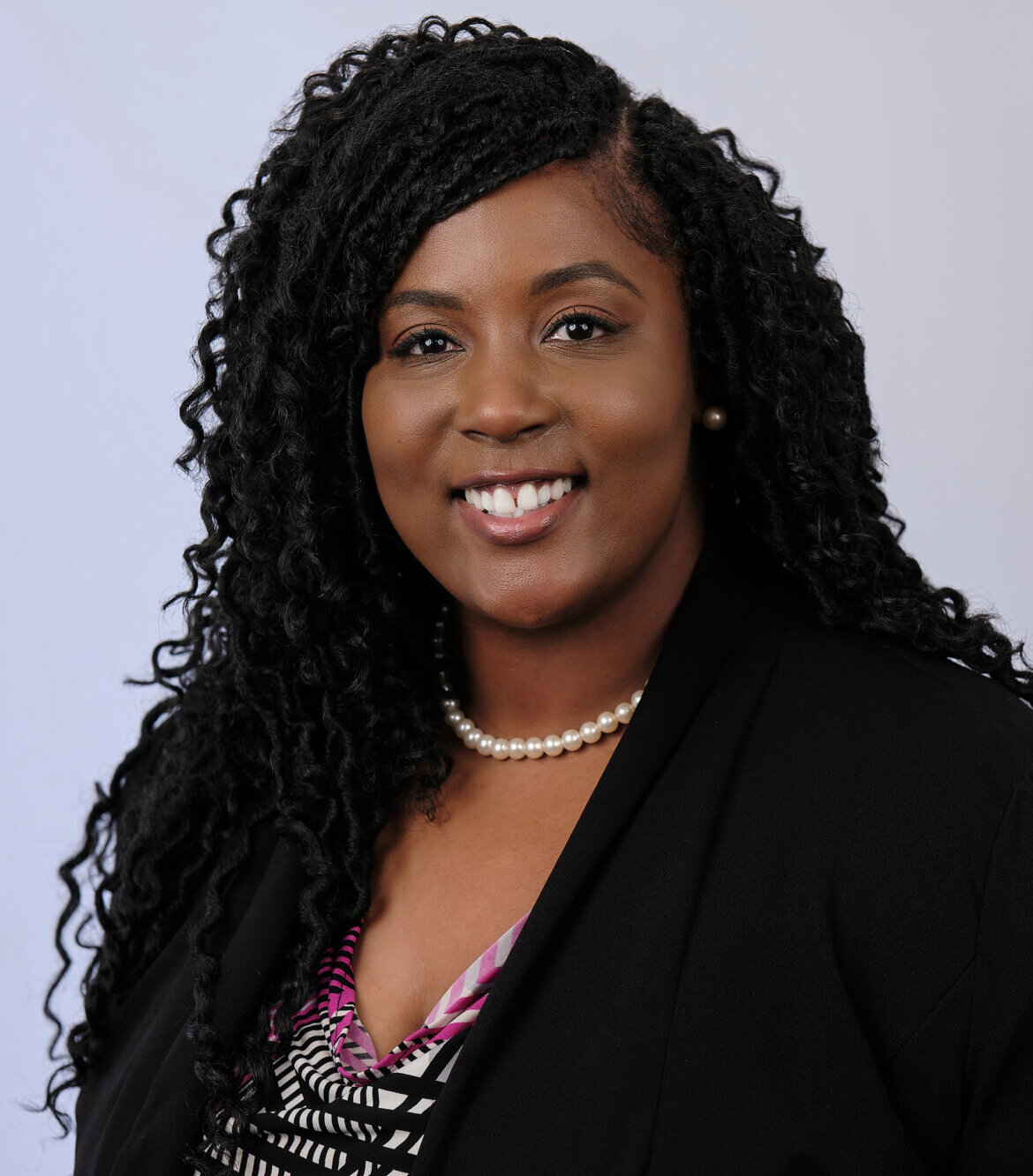 A woman with long curly hair smiles at the camera. She wears a black blazer, a patterned blouse, a pearl necklace, and stud earrings, with a light gray background.
