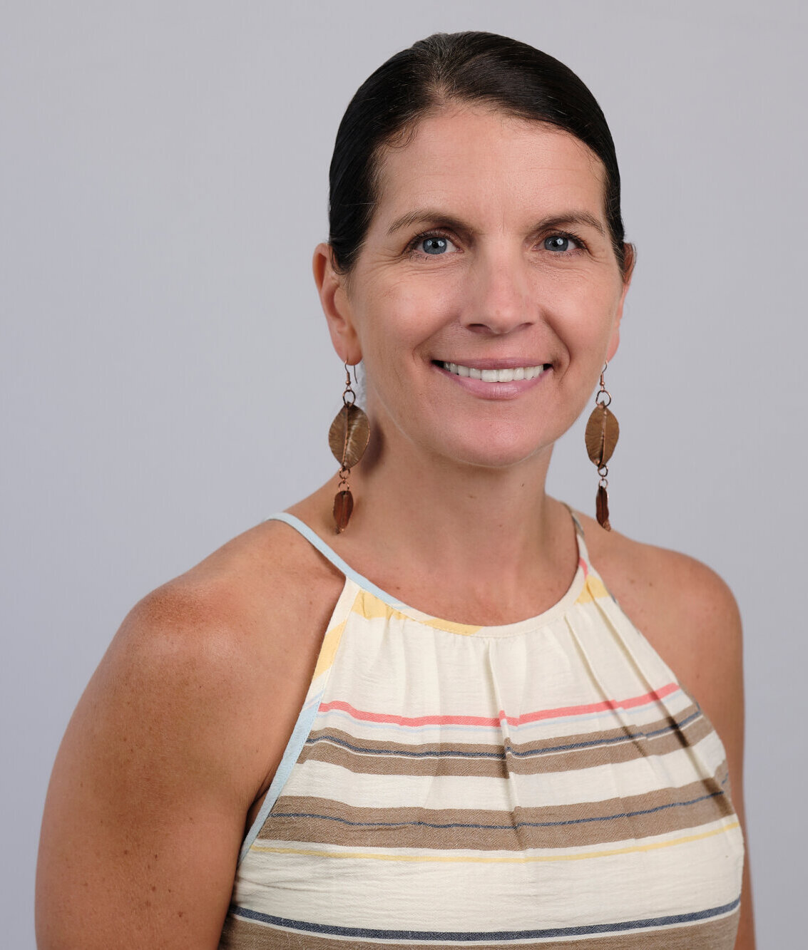 A woman with dark hair tied back, wearing dangling leaf-shaped earrings and a sleeveless striped top, smiles at the camera against a plain light gray background.