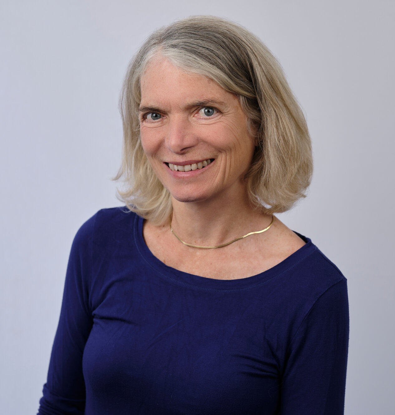 A woman with shoulder-length gray-blonde hair smiles at the camera. She is wearing a dark blue top and a thin gold necklace, standing against a plain light-colored background.