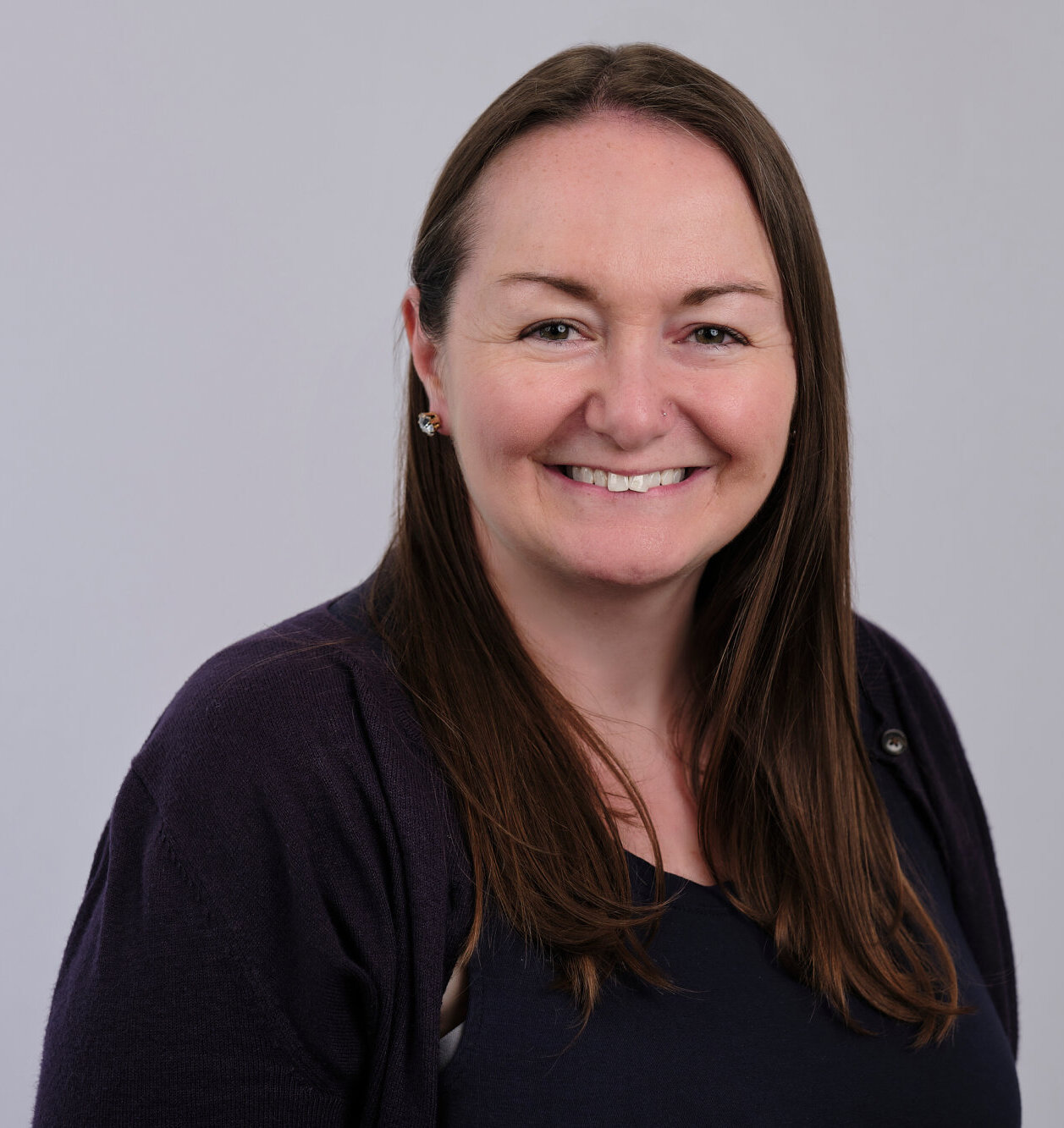 A woman with long brown hair, wearing a dark top and a dark cardigan, smiles at the camera against a plain light gray background.
