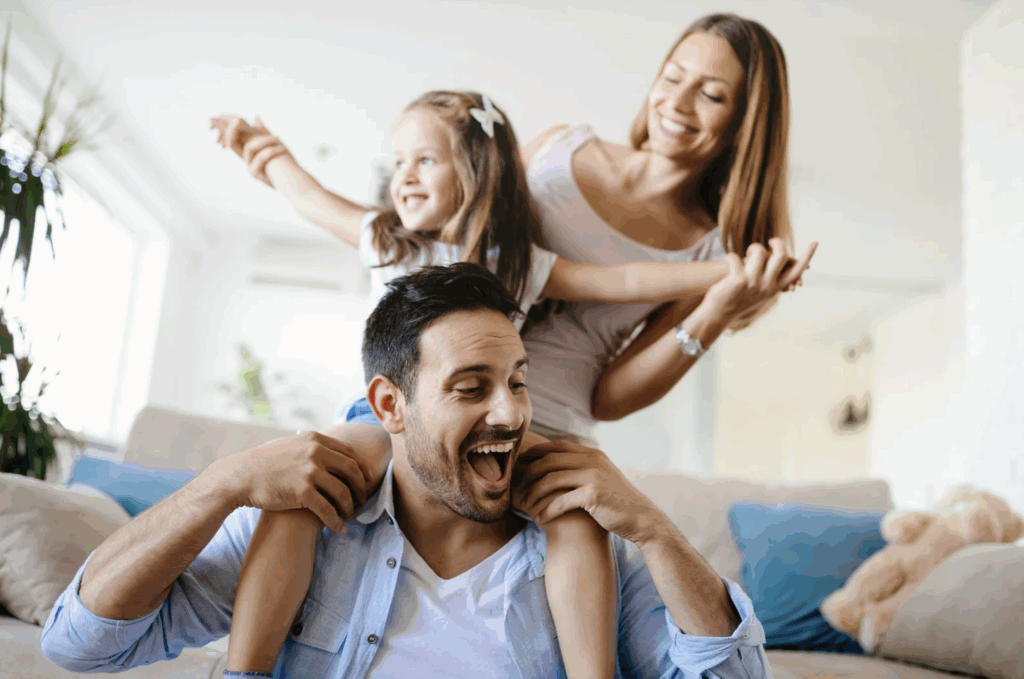 A cheerful family at home: a smiling man sits on a couch with a young girl on his shoulders, while a woman stands behind them, all laughing and enjoying time together in a bright living room—a perfect setting for research on happiness.