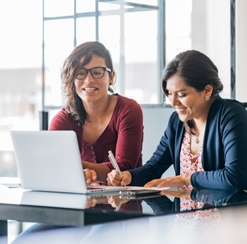 Two women sit at a table working together on a laptop. One woman is typing while the other is writing with a pen and smiling. They appear to be collaborating in a bright, modern office space.