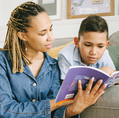 A woman and a young boy sit on a couch reading a book together. The woman holds the book open and reads aloud while the boy follows along, looking at the pages intently.