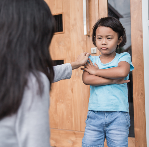 A young child stands outside with arms crossed, looking upset. An adult, seen from behind, gently touches the child’s shoulder in a comforting gesture near a wooden door.
