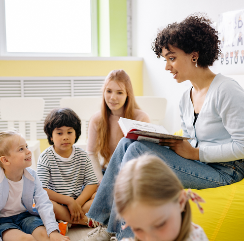 A woman reads a book to a group of young children sitting on the floor in a bright classroom, while another adult sits nearby, watching and smiling.