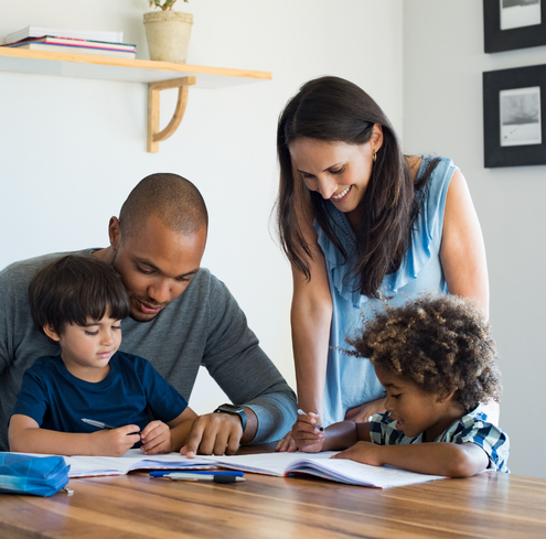 A man and a woman help two young children with homework at a wooden table, all smiling and engaged in the activity in a bright, homey room.