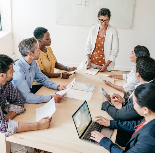 A group of six people sit around a conference table with documents and laptops as a woman standing at the head of the table leads a discussion, smiling.