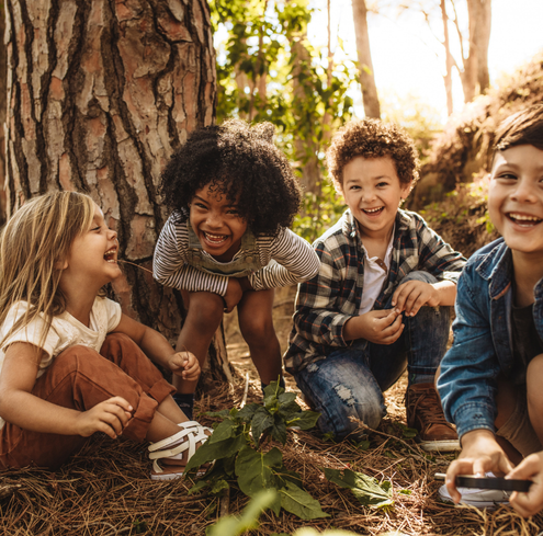 Four young children are laughing and playing together outdoors under a large tree on a sunny day. They are surrounded by pine needles and greenery, enjoying each others company and having fun in nature.