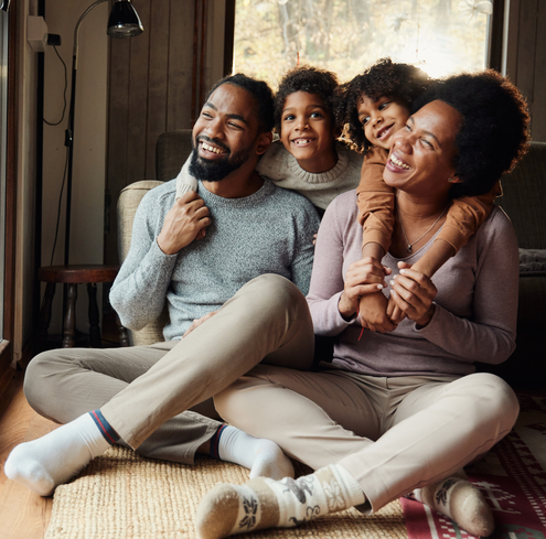 A smiling family of four sits on the floor by a window. Two adults sit cross-legged at the front, while two children lean over their shoulders from behind, all laughing together in a cozy, sunlit room.
