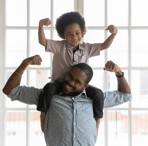 A smiling man carries a cheerful child on his shoulders. Both are flexing their arms in a playful pose, standing in front of large, bright windows.