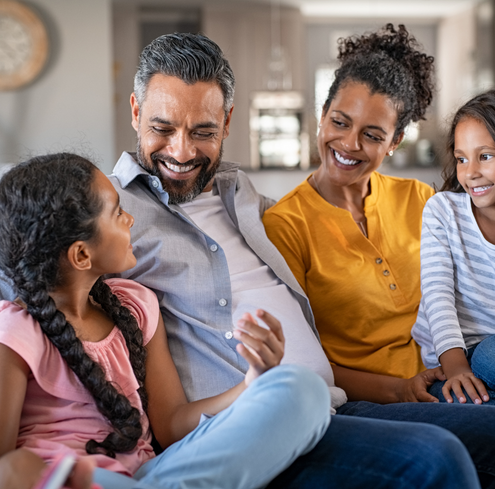 A family of four sits together on a couch, smiling and talking. The father and mother are in the middle, with two young daughters on either side, all appearing happy and engaged in conversation.