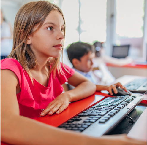 A young girl wearing a pink shirt uses a computer in a classroom, with another student in the background, both focused on their screens.