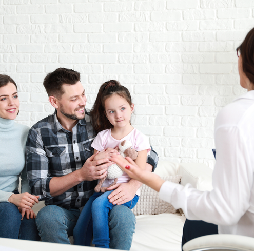 A smiling couple and their young daughter sit on a couch, talking with a woman in a white shirt who faces them, gesturing as she speaks. The girl holds a stuffed animal in her lap.