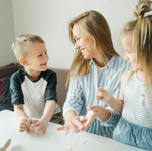 A smiling woman sits at a table with a young boy and girl, all laughing and playing with flour while baking together in a bright room, turning the fun into a playful training session for little chefs.
