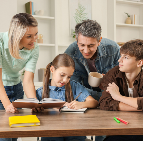 A woman and a man provide training as they help two children with homework at a table. The girl writes in a notebook while the boy listens. Books, pens, and a cup are on the table in a bright, cozy room.