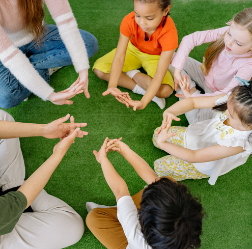 Five children and two adults sit in a circle on green carpet, touching fingers together, possibly playing a hand game or participating in a group training activity. Everyone appears engaged and focused.