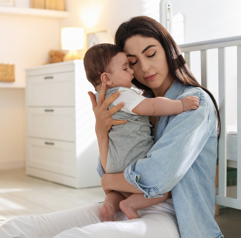 A woman sits on the floor of a nursery, gently holding and comforting a baby who rests their head on her shoulder. Sunlight streams in, creating a calm and nurturing atmosphere.