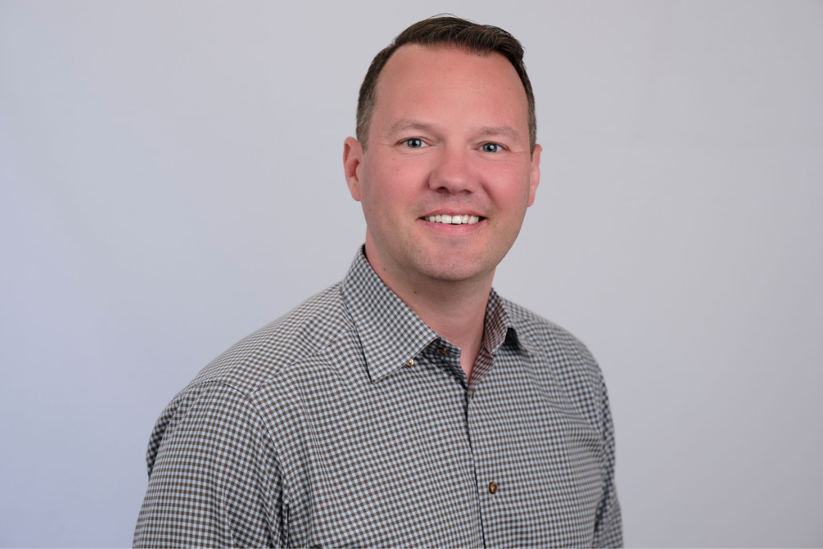 A man with short brown hair is smiling at the camera. He is wearing a checked button-up shirt and standing against a plain light gray background.
