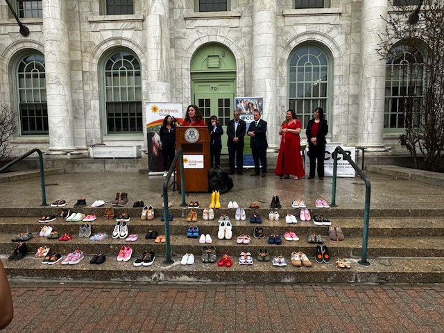 A speaker stands at a podium on courthouse steps in Berkshire County, flanked by others, with numerous pairs of shoes arranged for the Step Up for Kids event, symbolizing remembrance or awareness. Banners are displayed behind the group.