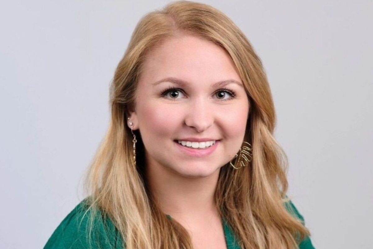 A smiling woman with long blonde hair wearing a green top and gold leaf-shaped earrings, posing in front of a plain light gray background.