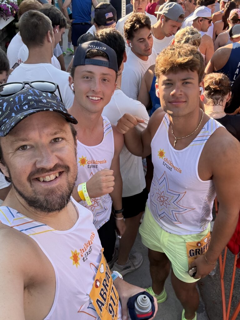 Three men in matching white Falmouth Road Race shirts and bibs smile for a selfie at the starting line of a crowded outdoor running event. Sunlight shines on their faces and other runners are visible in the background.