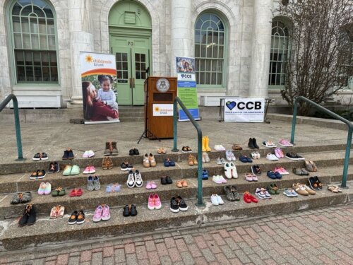 Several pairs of children's shoes are arranged on the steps outside a building with green doors, highlighting a Step Up for Kids event in Berkshire County. Signs from organizations such as Children's Trust and CCB are displayed near a podium at the top.