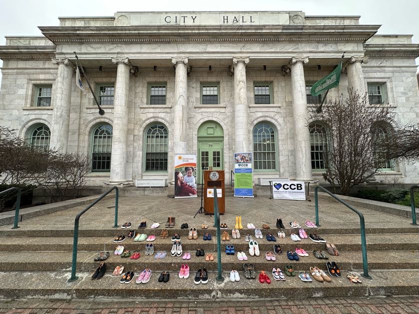 Rows of children's shoes are lined up on the steps outside the large, white city hall in Berkshire County. Banners and a podium hint at a Step Up for Kids event or memorial taking place by the entrance.
