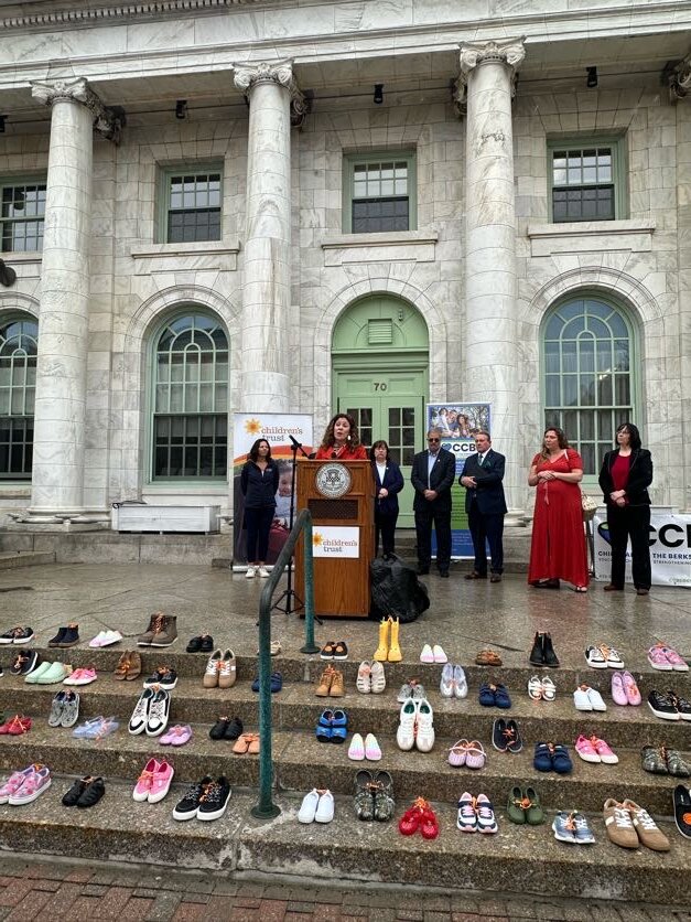 A woman speaks at a podium on the steps of a large building with columns, surrounded by others during a Berkshire County Step Up for Kids event. Numerous pairs of shoes, symbolizing kids advocacy, are arranged on the steps in front of her.