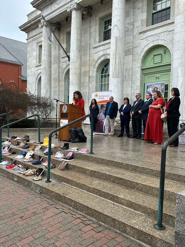A woman speaks at a podium on the steps of a large building with columns during a Step Up for Kids event in Berkshire County, as people stand behind her. Pairs of shoes and banners highlight the importance of this kids event.