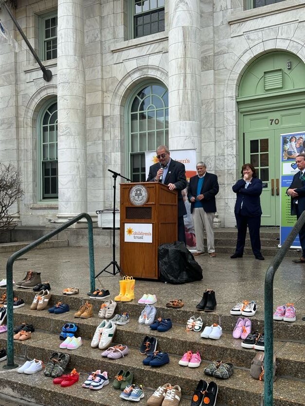 A man speaks at a podium on the steps of a building with tall columns during the Step Up for Kids event in Berkshire County, while dozens of children's shoes are arranged on the steps and several others stand nearby, listening.