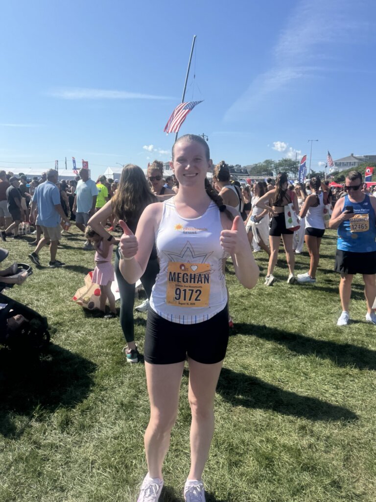 A woman wearing a race bib and athletic gear stands on grass, smiling and giving two thumbs up at an outdoor event with a crowd and American flag in the background on a sunny day, celebrating her accomplishment at the Falmouth Road Race.