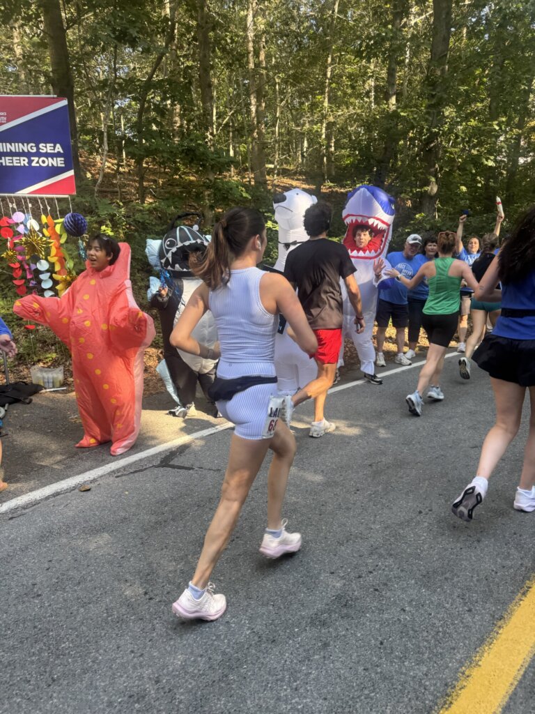 Runners pass a group in sea creature costumes, including a pink starfish and a blue shark, in a wooded area during the Falmouth Road Race. A sign reads CHEERING SEA CHEER ZONE.
