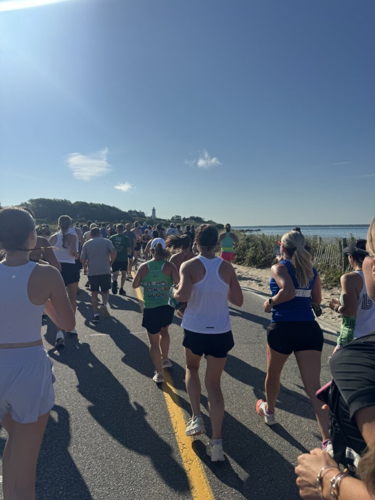A group of runners participate in the Falmouth Road Race on a sunny day, with waterfront views and a lighthouse visible in the background.