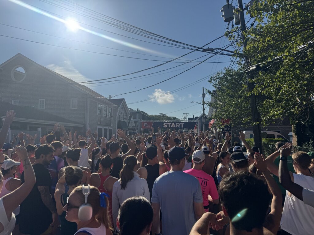 A crowd of runners raises their hands at a sunny outdoor Falmouth Road Race starting line, surrounded by houses and trees, with a START banner visible in the background.