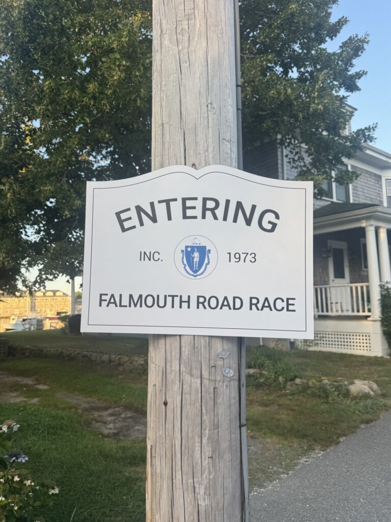 A white sign on a utility pole reads Entering Falmouth Road Race Inc. 1973 with a Massachusetts state seal, marking the proud heritage of the renowned Falmouth Road Race, in front of a house and trees.