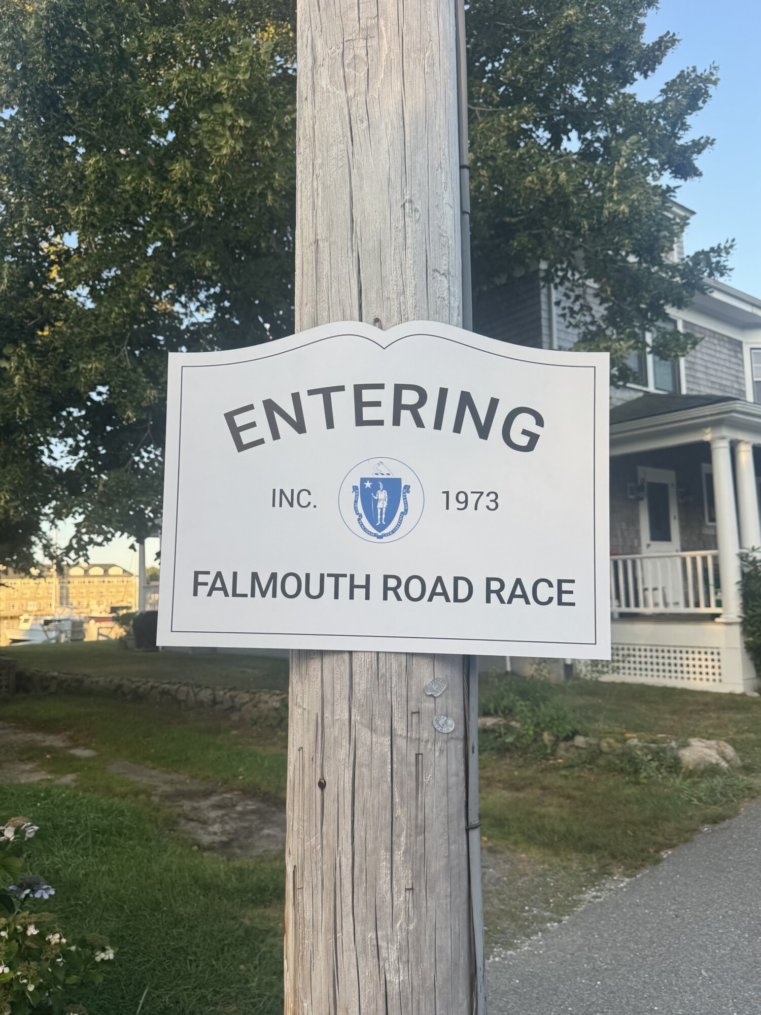 A white sign on a utility pole reads Entering Falmouth Road Race Inc. 1973 with a Massachusetts state seal, marking the proud heritage of the renowned Falmouth Road Race, in front of a house and trees.