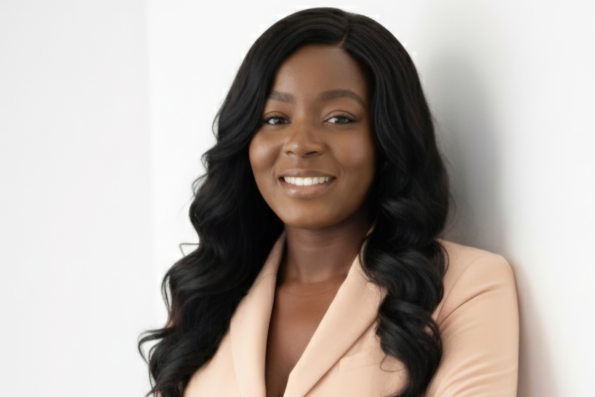 A woman with long, wavy black hair wearing a light beige blazer smiles confidently while leaning against a white wall.