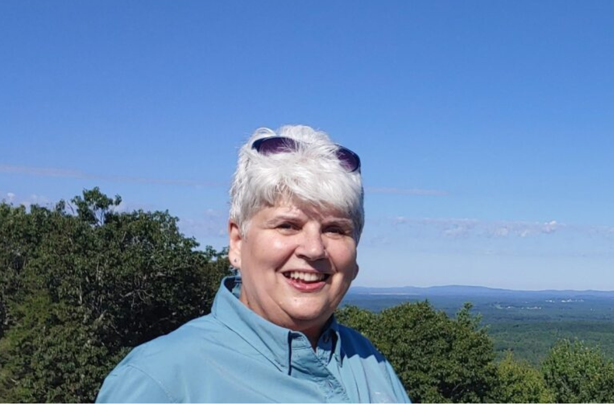 A smiling older woman with short white hair and sunglasses on her head stands outdoors with green trees and a distant blue landscape under a clear sky.