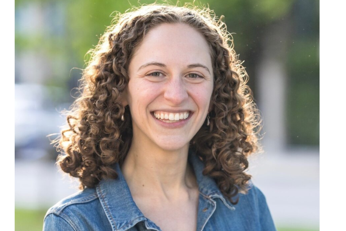 A woman with curly brown hair and a wide smile stands outdoors, wearing a blue denim jacket. The background is blurred with green trees and sunlight.