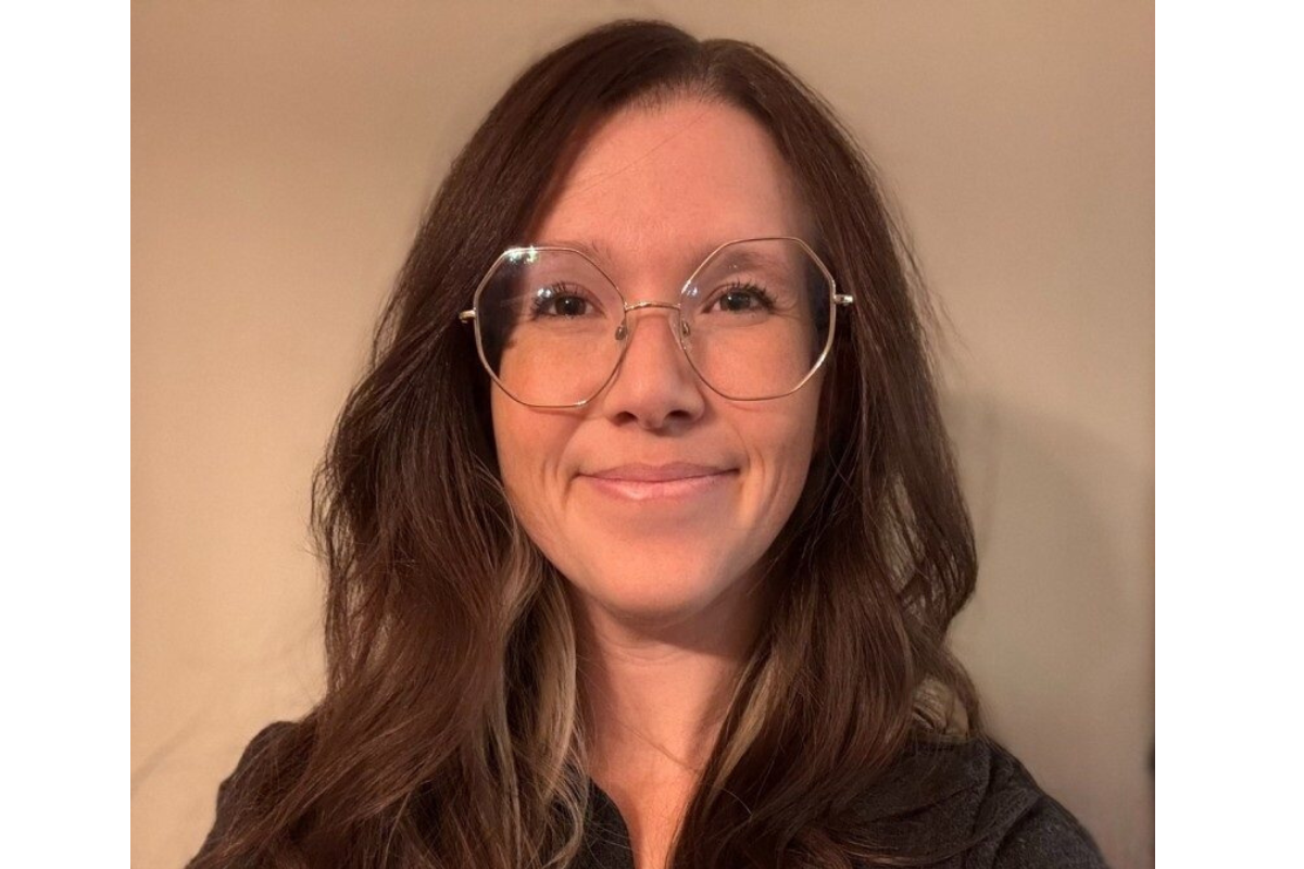 A woman with long brown hair and large, round glasses smiles softly at the camera. She is wearing a dark top and is posed in front of a plain beige background.