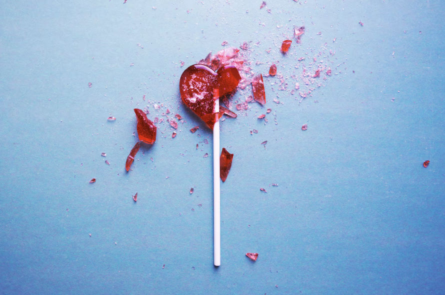 A red heart-shaped lollipop on a stick is broken, with scattered candy shards on a light blue background.