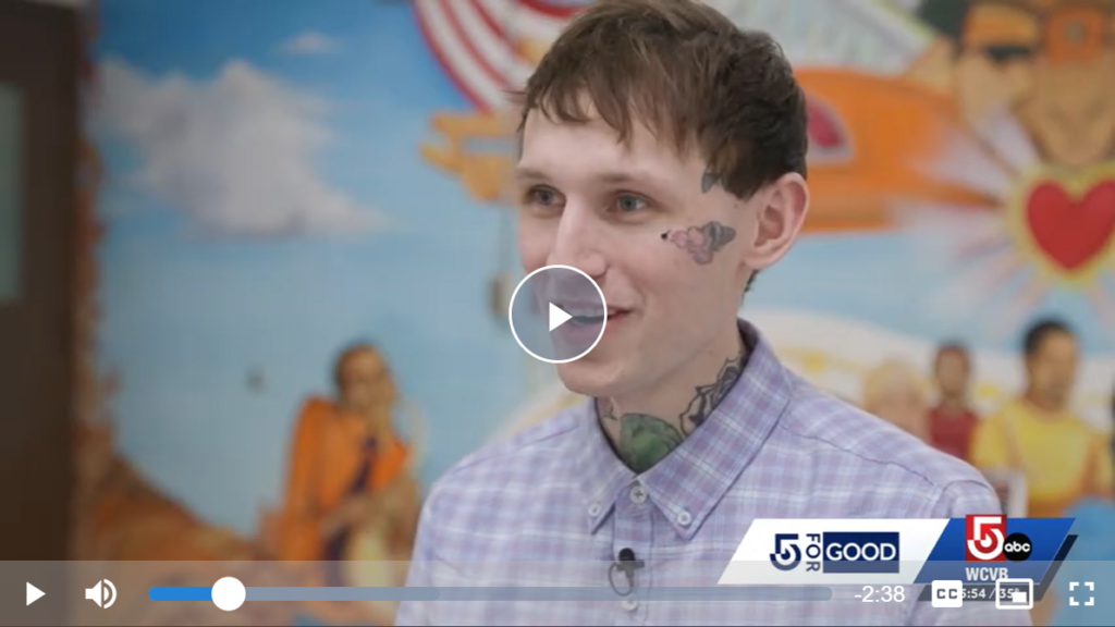 A young person with tattoos on their face and neck smiles while standing in front of a colorful mural. They are wearing a light purple plaid shirt. Video player controls and news station logos are visible on the screen.