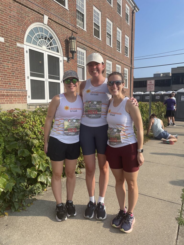 Three people stand smiling outside in athletic wear and matching “Children’s Trust” shirts, ready for the Falmouth Road Race. A brick building with tall windows, greenery, and a sidewalk are visible in the background on a sunny day.