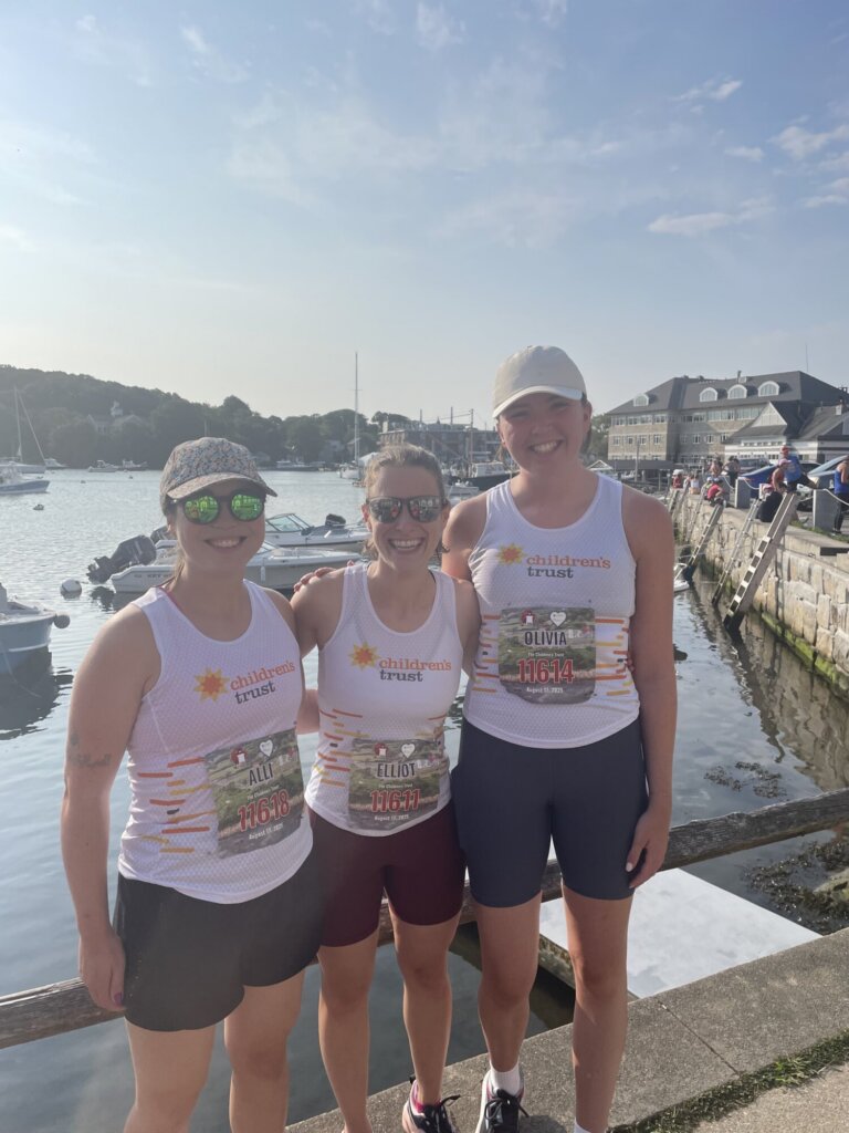 Three women in matching athletic gear and race bibs stand smiling by a marina, with boats and buildings in the background on a sunny day, celebrating their finish at the Falmouth Road Race.