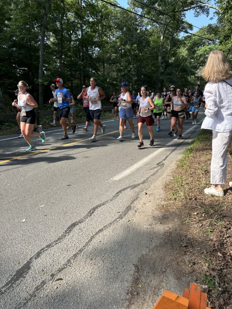 A group of runners participating in the Falmouth Road Race move along a tree-lined road, while a woman in light-colored clothing stands on the grass at the roadside, observing as sunlight filters through the trees.